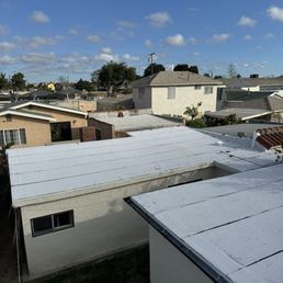 The image shows a view of several residential buildings with flat and sloped roofs, some covered with white roofing material. The houses are closely spaced, with trees and utility poles visible in the background. The sky is mostly clear with a few scattered clouds, indicating a sunny day. The scene appears to be in a suburban area with typical single-family homes.