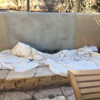 Always Construction Services - a patio with a blanket on it - The image shows a stone platform with a white fabric draped over it, positioned outdoors near a concrete wall. The fabric appears to be a sheet or cloth, arranged in a haphazard manner, possibly serving as a makeshift bed or covering. A wooden chair is partially visible in the foreground, and the background includes trees and natural scenery, indicating an outdoor setting. - Stucco Services Near Me - Santa Fe, New Mexico