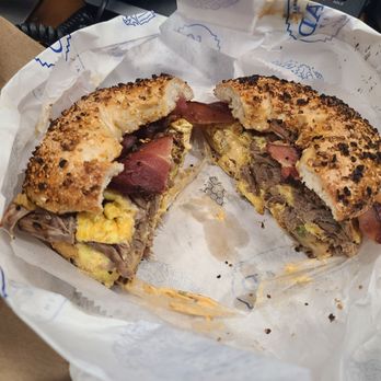 Spread Bagelry - A bagel sandwich with a toasted, seed-covered bagel, filled with egg, meat, and other ingredients, cut in half to reveal the fillings. The sandwich is placed on branded paper inside a paper bag, with some crumbs and sauce visible. - Bagels Near Me - Dresher, Pennsylvania