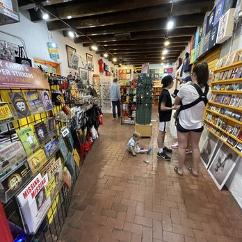 Guerrilla Graphix - Inside a retail store, several customers, including a small child sitting on the floor, browse art cards, stickers, and framed prints. The store has shelves filled with various merchandise, including art supplies and decorative items. The ceiling features exposed wooden beams, and the floor is made of brick tiles. The store appears to sell art-related products and collectibles. - Printing Services Near Me - Albuquerque, New Mexico