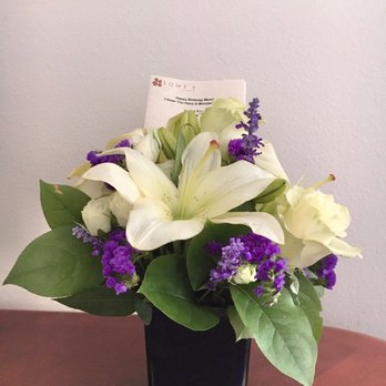 Lowe's Flowers - Arrangement my mother received - A floral arrangement includes white lilies, roses, and purple statice flowers, arranged with green leaves in a black square vase. A note from Lowe's Flowers is inserted behind the flowers, partially visible. The bouquet is placed on a wooden surface against a plain, light-colored wall. - Florists Near Me - Marshalltown, Iowa