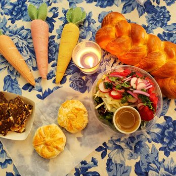 Ruthie's Bagels - Chocolate Toffee Matzah w/pistachios, challah, potato knishes, Shaved fennel, radishes, strawberries, greens & pomegranate vinaigrette - The image shows a table covered with a blue floral tablecloth, displaying a bowl of mixed salad with radishes, strawberries, and greens, along with two croissants, a plate of chocolate bark with nuts, and three plush carrot toys in pastel colors. A lit candle adds ambient lighting to the scene. - Bagels Near Me - Albuquerque, New Mexico