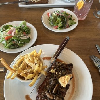 Devil's Kitchen - The image shows a dining scene with a main plate featuring a cooked steak topped with sautéed mushrooms and a lemon wedge, accompanied by a side of French fries. Two salads with mixed greens, tomatoes, and onions are also visible on separate plates. In the background, there is a glass of pink lemonade with a lemon slice, a bowl of French fries, and a dessert with a fork and knife on the table. The setting appears to be a restaurant with a wooden table surface. - New American Near Me - Grand Junction, Colorado