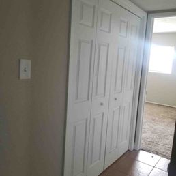 The image shows a white, double-door closet with paneled doors, situated against a beige wall. The closet is located in a room with brown tiled flooring. To the right, there is an open doorway leading to another room with carpeted flooring and a window that allows natural light to enter. The overall setting appears to be a residential interior space, possibly a bedroom or hallway.