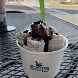 Cookies and cream rolled ice cream with chocolate syrup - A cup of cookies and cream rolled ice cream is shown, topped with chocolate syrup and a green spoon. The ice cream is arranged in rolled form inside a white cup with a Frozen Boba logo. The cup is placed on a black metal table, with a background of a parking lot and a building visible through a window.