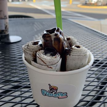 Frozen Boba - Cookies and cream rolled ice cream with chocolate syrup - A cup of cookies and cream rolled ice cream is shown, topped with chocolate syrup and a green spoon. The ice cream is arranged in rolled form inside a white cup with a Frozen Boba logo. The cup is placed on a black metal table, with a background of a parking lot and a building visible through a window. - Ice Cream & Frozen Yogurt Near Me - Evesham Township, New Jersey
