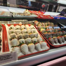 The image shows a glass display case containing trays of chocolate-covered strawberries. The strawberries are decorated with various chocolate drizzles and coatings, arranged neatly on white paper-lined trays. A label indicates one variety as 'Chocolate Covered Strawberry.' The display is part of a dessert shop offering chocolate treats, with packaging and other chocolates visible in the background.