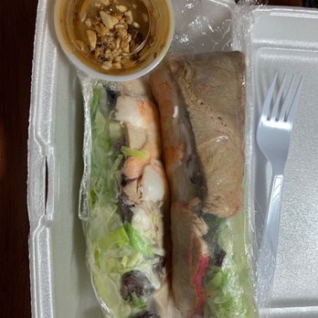 Bamboo by Kulantro - Spring Mix Blend - Two Vietnamese spring rolls wrapped in clear plastic are placed on a foam tray. One roll contains shrimp, lettuce, and herbs, while the other appears to contain grilled meat and vegetables. A small plastic container of dipping sauce with crushed peanuts is also on the tray. The tray is on a wooden surface, and a plastic fork is positioned to the right of the tray. - Vietnamese Near Me - Albuquerque, New Mexico