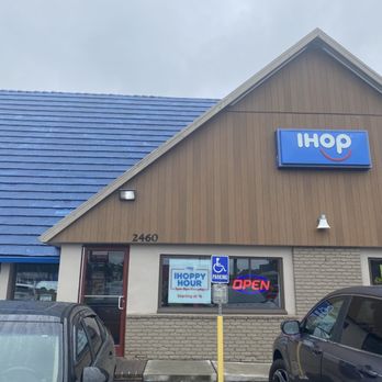 IHOP - Outside - The photo depicts the exterior of an IHOP restaurant located at 2460, featuring a brown and blue building with a sloped roof. There are parked cars in front, including a black vehicle and a gray one. A handicap parking sign is visible, along with a window displaying an IHOP Hour sign and an illuminated 'OPEN' sign. The sky above is overcast. - Breakfast & Brunch Near Me - Union, New Jersey
