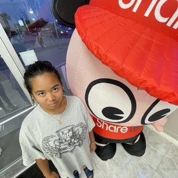 Sharetea - A girl with dark hair tied back, wearing a gray T-shirt with a graphic design, stands indoors next to a Sharetea mascot costume. - Bubble Tea Near Me - Palmdale, California
