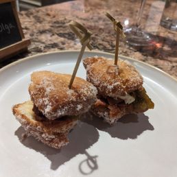 Cheese course: cheese inside of beignets - Two beignet-style appetizers with skewers are placed on a white plate. They are dusted with powdered sugar and contain fillings, possibly cheese and vegetables. The plate is on a granite countertop, with a small chalkboard sign and glasses visible in the background.