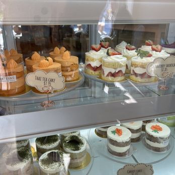 Maprang Bakery & Cafe - The image features a glass display case containing a selection of cakes and desserts at Maprang Bakery & Cafe. Visible items include Thai tea cakes, strawberry shortcake, matcha red bean cake, and carrot cake, each with decorative toppings and layered presentation. Price labels indicate the cost of each item, and the case is illuminated to highlight the desserts. The background shows baked goods on an upper shelf, with reflections on the glass surface. - Desserts Near Me - Somerville, Massachusetts