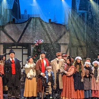 Old Town Temecula - Townspeople standing together in the snow on Christmas morning - A group of actors dressed in historical costumes perform on stage, with a set resembling a village scene and snow falling. - Local Flavor Near Me - Temecula, California