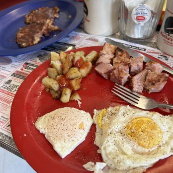 Martha's Diner - Eggs and things. - A breakfast plate featuring fried eggs, diced potatoes with ketchup, and pork chunks, with a coffee mug and condiments in the background. - American Near Me - Ellsworth, Maine