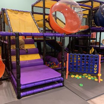 Wisco Kidz Place - The image shows an indoor children's play area with a multi-level soft play structure. It includes purple and yellow padded steps, a large orange tube slide, and a ball pit filled with multicolored balls. Adjacent to the structure is a Connect Four game with green and yellow discs. The flooring is covered with safety mats, and the background has a light-colored wall and metal support beams. - Playgrounds Near Me - Madison, Wisconsin