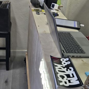 Discovery Records - Check out! - The image shows a reception desk with a silver laptop, a smartphone, a small camera, and various electronic accessories such as a speaker and chargers. The desk is made of light-colored wood, and there is a white wall behind it. A black chair is partially visible to the left, and a small stack of magazines or flyers is on the desk. The setting appears to be an office or retail space. - Vinyl Records Near Me - Temecula, California