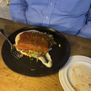 Vue - Bison meatloaf - A person in a blue button-up shirt is sitting at a wooden table in a restaurant. In front of them is a black plate with a serving of bison meatloaf topped with sauce and garnished with herbs. The plate rests on a wooden table, and a fork is placed on the side. An empty white plate with some sauce remnants is also visible on the table. - Cocktail Bars Near Me - Wadsworth, Ohio