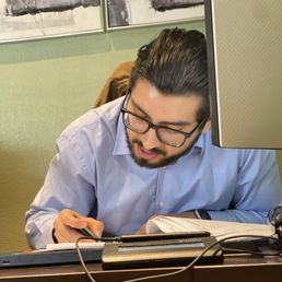 That's the man!! - A man with glasses and dark, slicked-back hair is seated at a desk, writing in a notebook with a pen, surrounded by office equipment.
