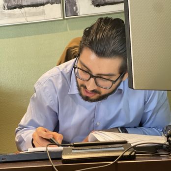 MacTax Solutions - That's the man!! - A man with glasses and dark, slicked-back hair is seated at a desk, writing in a notebook with a pen, surrounded by office equipment. - Tax Services Near Me - Downey, California