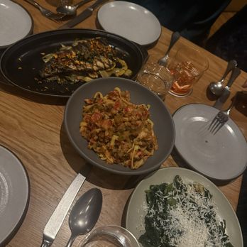 Tavernetta - Tagliatelle, Rapini, and Branzino - The image shows a wooden dining table set with three main dishes of Italian cuisine: a fish fillet garnished with herbs on a black platter, a bowl of pasta with a tomato-based sauce, and a plate of cooked greens topped with grated cheese. There are also glasses of water, small condiment containers, and empty plates with forks and knives arranged around the table, indicating a meal setting. - Italian Near Me - Denver, Colorado