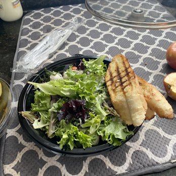 Nick's Pizza & Pub - Tomato Mozzarella Salad - A black plastic plate contains a mixed green salad with different types of lettuce, including green and purple leaves. Two slices of grilled bread with grill marks are also on the plate. The plate is on a patterned placemat with a geometric design, on a dark countertop. Nearby, there are other food items and utensils, including a wrapped utensil and a glass cover. - Pizza Near Me - Elgin, Illinois