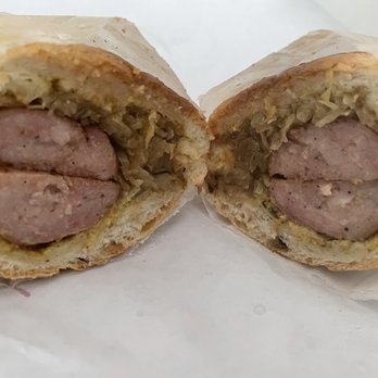 Polka Deli - Kielbasa sandwich - A sandwich featuring a cooked kielbasa sausage and sautéed cabbage inside a soft bread roll, cut in half to reveal the filling. The sandwich is placed on a white surface with some paper wrapping visible. - Polish Near Me - Silver Spring, Maryland