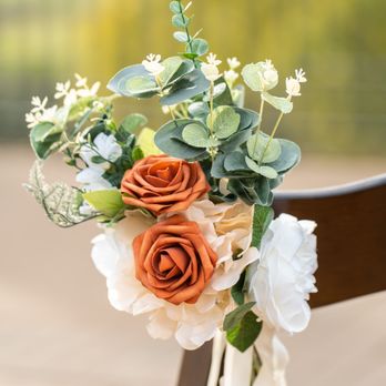 TDE Wedding - A floral decoration featuring two orange roses, white flowers, and eucalyptus leaves is attached to a wooden chair. The arrangement is tied with a white ribbon, and the background shows a blurred outdoor scene with green and yellow hues, suggesting an outdoor event setting such as a wedding. - Wedding Planning Near Me - Millbrae, California