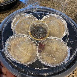 Mala Spicy Soup Dumplings (4 Pcs) / 麻辣烫包 - Four soup dumplings are arranged in a clear plastic container with a small cup of soy sauce in the center, on a granite countertop.