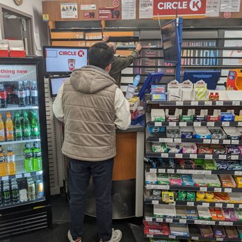 Circle K - Payment area - A man stands at the checkout counter of a Circle K, with a cashier assisting him. The store has snack shelves and a beverage fridge nearby. - Convenience Stores Near Me - Aurora, Ontario