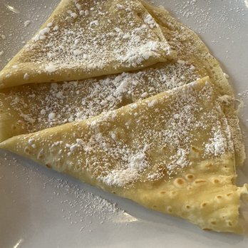 Elevate Coffee - Lemon Crepe - Two folded crepes dusted with powdered sugar are presented on a white, square plate. The crepes are golden-brown and folded into triangles, with powdered sugar evenly sprinkled on top. The plate is placed on a wooden surface, suggesting a dining setting. The image emphasizes the dessert's appearance and presentation, suitable for a cafe or breakfast menu. - Coffee & Tea Near Me - Phoenix, Arizona