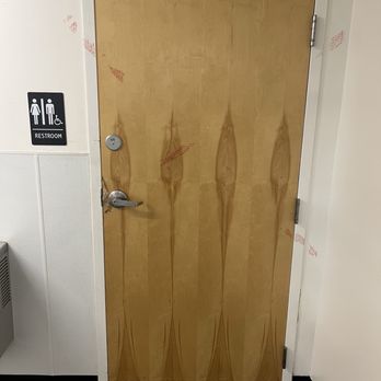 CVS Pharmacy - A wooden door with a natural finish and visible wood grain patterns, equipped with a door closer at the top and a metal handle. The door is set within a white door frame and is adjacent to a wall with a restroom sign indicating accessible facilities for men and women. The surrounding area includes a section of a carpeted floor and a wall with a textured lower panel. - Drugstores Near Me - Peekskill, New York
