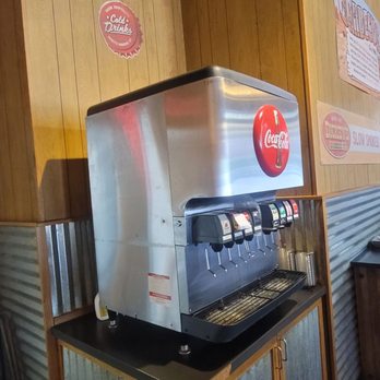 Dickey's Barbecue Pit - a hot dog machine - A stainless steel Coca-Cola soda fountain machine with multiple drink dispensers is positioned on a black countertop against a wooden-paneled wall. - Barbeque Near Me - Eagan, Minnesota