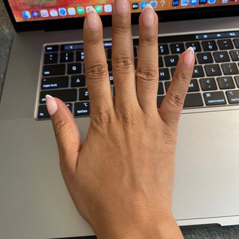 Omani  - Gel Polish (No-Chip) French Manicure - A hand with well-groomed, light pink, almond-shaped nails is resting on a laptop keyboard. The laptop screen shows a colorful background with app icons along the bottom. The setting appears to be a workspace or personal area. - Nail Salons Near Me - Chicago, Illinois