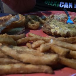 Chicken - fried Steak - A food plate featuring fried steak with gravy, French fries, onion rings, and additional food items, served on a pink plate. The setting appears to be casual dining, with a fork and a person partially visible in the background. The image is taken in low light, emphasizing the fried and savory foods.