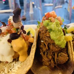 Fish Tacos - Two tacos with different fillings, topped with guacamole, salsa, and garnishes, served on a plate in a restaurant setting.