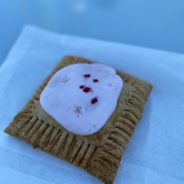 Raspberry Prickly Pear - A rectangular pastry with a golden-brown crust and ridged edges is topped with a layer of white frosting and small red sprinkles. The pastry is placed on a white paper napkin, with a blurred blue background.