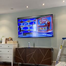 75 inch tv mounted to stucco wall. He only used 4 bolts and is on there incredibly strong! - A television is mounted on a light-colored wall, displaying a game show called 'Grocery Game.' Below the TV, a wooden cabinet with geometric patterns holds a water bottle on top. To the left, a white dresser contains towels and supplies. A ladder is positioned on the right side of the room, which has hardwood flooring and a patterned rug in the foreground. The setting appears to be an indoor space, possibly a lounge or waiting area.