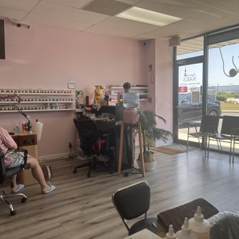 Super Star Nails - Interior - The image shows the interior of a nail salon with a pink wall, a desk with nail supplies, and a woman seated in a black chair. There are multiple chairs along the window, which offers a view of parked cars and a building outside. The space includes a water dispenser, a potted plant, and various nail polish bottles and tools. The ceiling has fluorescent lighting, and the floor is covered with wood-patterned vinyl. - Nail Salons Near Me - Frankfort, Illinois