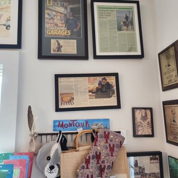 Bennett Shellfish - Wall features multiple framed newspaper articles, photographs, and awards, arranged in a gallery style. A shelf below holds a sign reading 'Montauk' and various items such as a crocheted animal face, a bag, and a fabric with fish illustrations. The setting suggests a personal or business space decorated with memorabilia and recognition related to Bennett Shellfish in Montauk, NY. - Seafood Near Me - Montauk, New York