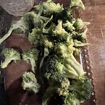 Uncommon Ground - Broccoli. Which was half the salad - Multiple broccoli florets are arranged on a wooden table, with some florets overlapping each other. The image includes a clear plastic container in the upper left corner, and the surface appears to be a dark wooden table. The lighting is dim, and the focus is on the broccoli pieces, which are fresh and green. - Breakfast & Brunch Near Me - Chicago, Illinois