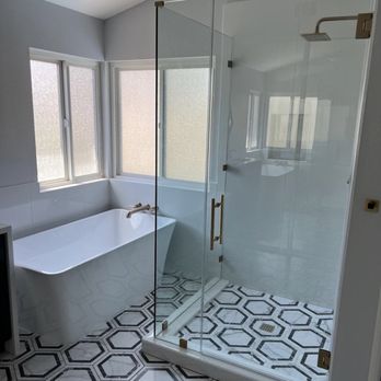 Cova Construction - Shower and tub now - Bathroom featuring a white bathtub near frosted windows and a glass shower with a brass handle, set on black and white patterned tile flooring. - General Contractors Near Me - Corona, California