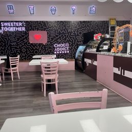 Cute decor - The image shows the interior of a dessert shop with a black and pink color scheme. Pink chairs are arranged around white tables on a wood-look floor. The back wall has neon signs, including ice cream icons, and phrases such as 'Sweeten Together' and 'Choco Addict.' There is a counter with a display case and a ladder on the right side. The ceiling has decorative white cloud-like elements.