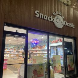 The cutest mom and pop shop owned by grandparents. Very clean and fully stocked on all the Asian snacks, icecream, candies, and drinks. - Snack Oasis, a convenience store with a wooden facade, features a glass entrance with an illuminated 'OPEN' sign and colorful posters on the door.