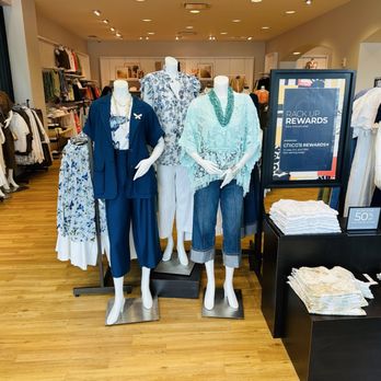 Chico's - Women's Apparel. - Mannequins dressed in women's clothing, including tops, pants, and accessories, are positioned at the entrance of Chico's store in Boca Raton, FL. The display features casual and stylish outfits, with a sign promoting rewards and discounts. The store interior shows racks of clothing, shelves with folded items, and a warm, well-lit environment with wooden flooring. - Women's Clothing Near Me - Boca Raton, Florida