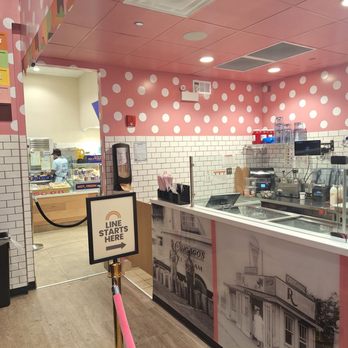 The Original Rainbow Cone - The image shows the interior of an ice cream shop with pink walls decorated with white polka dots and a white tiled lower wall. The counter area includes ice cream machines, cups, and utensils. A sign indicates where to line up, and a doorway leads to the kitchen or service area where staff are visible. The ceiling has recessed lighting and ventilation panels, creating a bright and clean environment. - Ice Cream & Frozen Yogurt Near Me - Chicago, Illinois