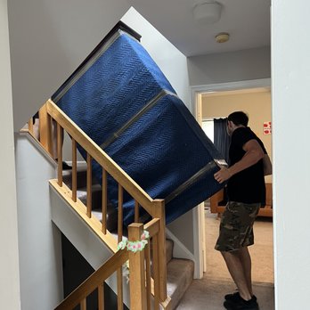 ZMax Movers - They definitely knew what they were doing! - A man wearing a black shirt and camouflage shorts is lifting a large blue mattress with a quilted pattern up a staircase inside a residential house. The staircase has wooden railings decorated with a small floral garland. The interior features beige carpeting, white walls, and a doorway leading to a living room with furniture and curtains. The scene captures the process of moving furniture within a home. - Movers Near Me - Fairfax, Virginia