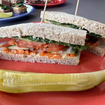 Great Harvest Bread - Hummus sandwich - A sandwich with hummus, tomato, cucumber, and leafy greens on white bread, served with a pickle on a pink plate. - Coffee & Tea Near Me - Cedar Rapids, Iowa