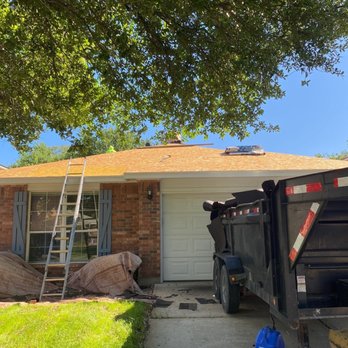 Cool Roofs - Full roof replacement - A single-story brick house with a white garage door and a window with shutters is shown. A ladder is positioned against the roof, which appears to be under repair or maintenance. A trailer is parked in the driveway, and various tools and equipment are scattered on the ground, suggesting ongoing roofing work. The scene is shaded by large trees, with a clear blue sky overhead. - Roofing Near Me - Austin, Texas