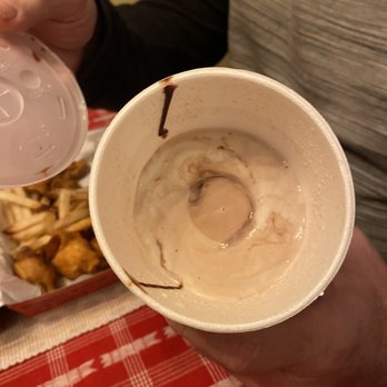 Slim Chickens - A "large" shake. What's wrong with this pic? - A disposable cup containing leftover ice cream and chocolate syrup, held by a person wearing a gray and black shirt. The cup shows streaks of chocolate syrup on the inside walls. In the background, a basket of fried food is visible on a red and white checkered tablecloth, indicating a casual dining setting. - Fast Food Near Me - Avon, Ohio