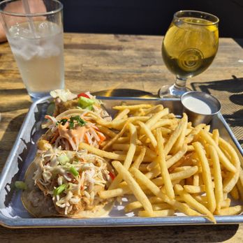 Commonwealth - Cauliflower Tacos. - A metal tray contains French fries, two tacos topped with vegetables and sauce, and a small cup of dipping sauce, placed on a wooden table. Two glasses of water and a glass of yellow beverage are also visible, with sunlight casting shadows on the table. The setting appears to be a casual dining restaurant. - Beer Bar Near Me - Modesto, California